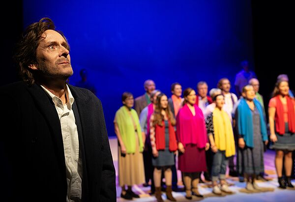 Ein Schauspieler auf der Bühne. Rechts im Hintergrund steht der Chor.