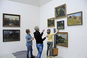 Drei Frauen stehen vor fünf Bildern an einer Wand. Eine der Frauen richtet das mittlere Bild aus.