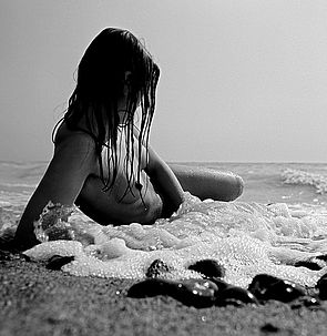 Eine nackte Frau liegt im seichten Wasser und wird von den Wellen umspült. Ihre langen Haare verdecken das Gesicht.