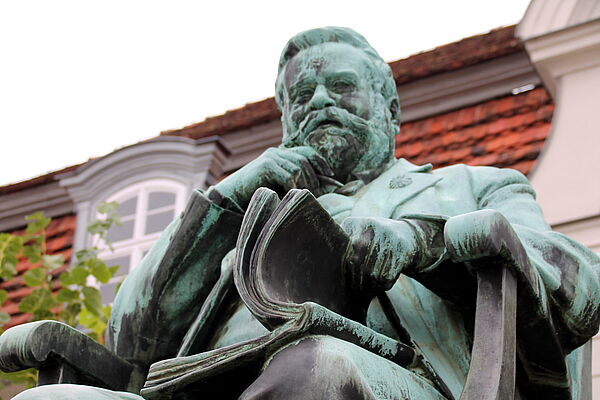 Fritz Reuter sitzt auf einem Stuhl. Eine Hand hat er ans Kinn geführt. Auf seinen Knien liegt ein Buch, das er mit einer Hand aufblättert. Hinter dem Denkmal steht das Literaturmuseum. 