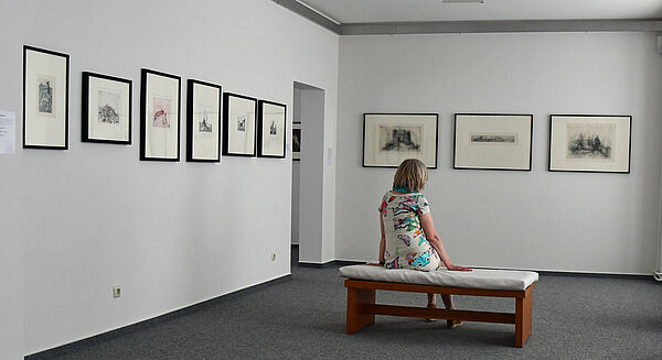 Eine Frau sitzt in einem Ausstellungsraum auf einer Bank, die mitten im Raum steht, und betrachtet Gemälde an der Wand.