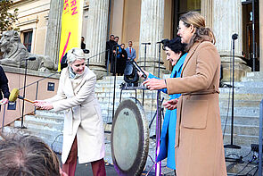 Drei Frauen vor dem Museum. Eine Frau schlägt den Gong zur Eröffnung. Die Frau ist Manuela Schwesig.