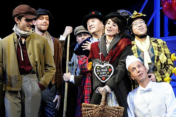 Sieben Schauspielerinnen und Schauspieler stehen in Kostümen nebeneinander und singen.