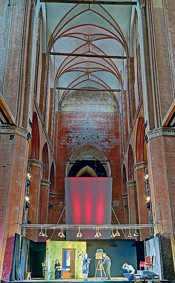Im Innenraum einer Kirche steht eine Bühne. Menschen stehen auf der Bühne und spielen Theater. 