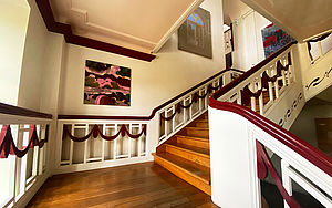Das Treppenhaus. Weißes Geländer mit rotem Handlauf. Holztreppen. Gelbe Wände. Ein Gemälde hängt an der Wand. 