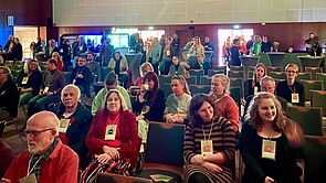 Ein Saal. Im Saal sitzen viele Menschen.