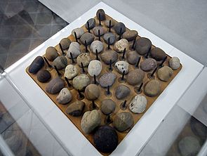 Auf einem Quadrat liegen in sechs Reihen jeweils sieben kleine Steine nebeneinander. Dazwischen befinden sich fünf Reihen mit jeweils sechs Nägeln.