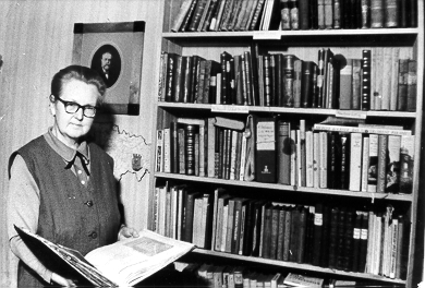 Eine Schwarz-Weiß-Aufnahme von Annalise Wagner. Sie steht mit einem aufgeschlagenen Buch in den Händen vor einem Bücherregal.