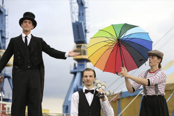 Drei Leute. Sie zeigen ein Kunststück. Eine Frau hält einen Regenschirm. Der Regenschirm ist bunt. 