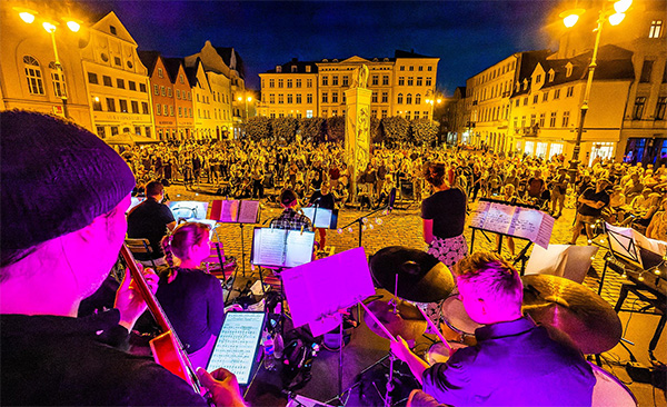 Ein großer Platz. Musiker spielen Musik. Viele Menschen schauen zu. Es ist dunkel. Der Platz ist beleuchtet.