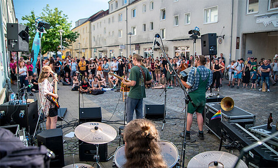 Eine Straße zwischen Häusern. Musiker spielen. Viele Leute schauen zu.
