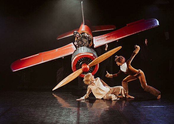 Eine Bühne. Auf der Bühne ist ein Flugzeug. Vor dem Flugzeug sind zwei Leute. Sie sind der kleine Prinz und ein Fuchs.